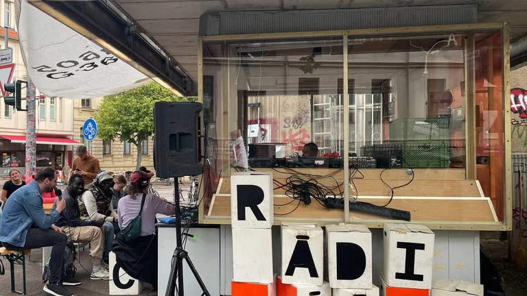 Kleiner mobiler Radiostand mit großen Buchstabenblöcken, die das Wort »RADIO« bilden, davor mehrere sitzende Personen auf Bänken.