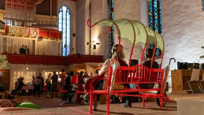 Two people are sitting on a red bench with a rose arch-like canopy in a church interior with people in the background.