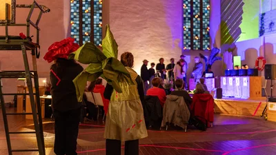 Personen mit großen Blumen-Kostümen stehen in einer Kirche vor einer Gruppe sitzender und stehender Menschen.