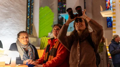 Three people indoors with colorful stained glass windows, one person holding binoculars pointed upward