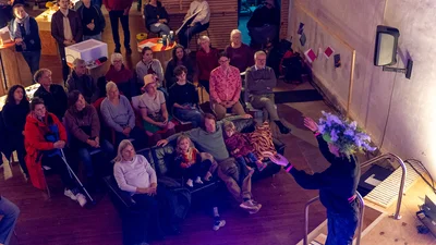 Person standing on a stage speaking to an audience seated on chairs and sofas