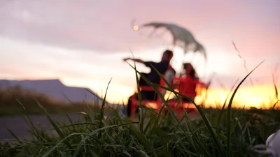 Blurred silhouettes of two people sitting on a bench in the grass, with a sunset in the background.
