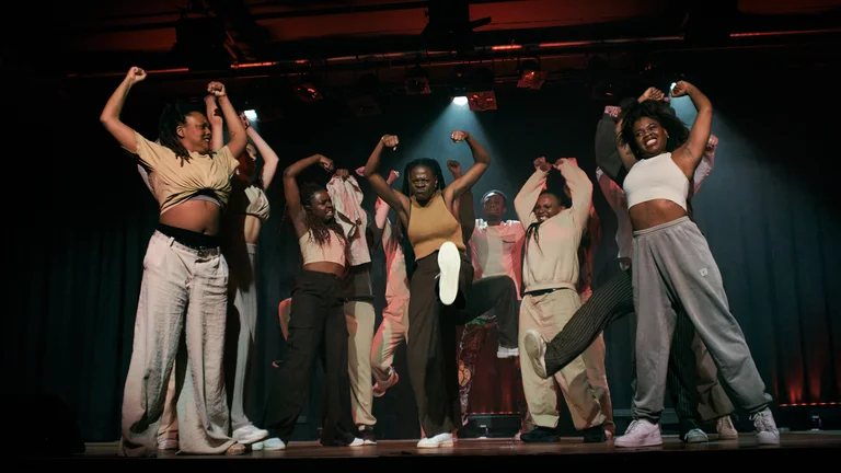 A group of young women dance on a stage, their arms stretched confidently upwards.