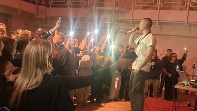 A young man is standing on a small stage, singing into a microphone, with an audience of young people in front of him.
