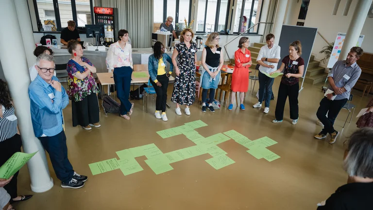 a group of people stand in a circle, with various pieces of paper lying on the floor in the centre