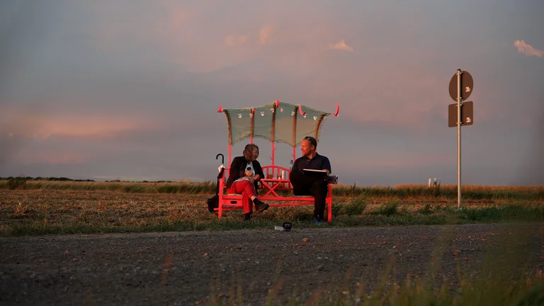 Two people are talking while sitting on a bench by a road in the countryside.