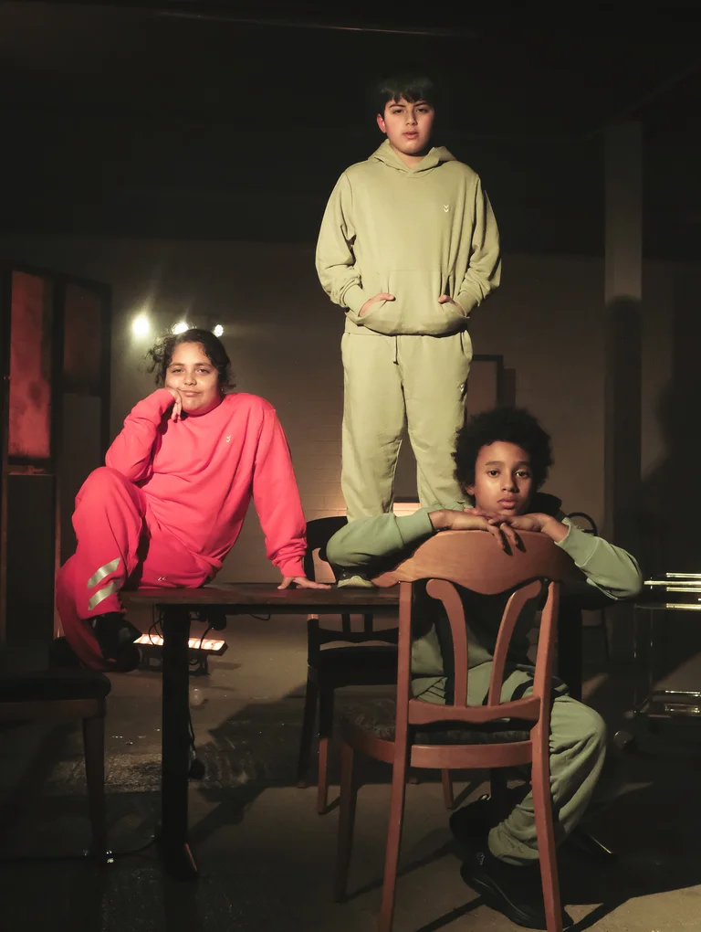 Three children are standing and sitting on a table and a chair, looking at the camera.