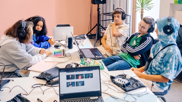 Ein Gruppe von Jugendlichen sitzt mit Kopfhörern um einen Tisch, auf dem verschiedene Technikgeräte wie Laptops, Mikrofone, Kabel liegen
