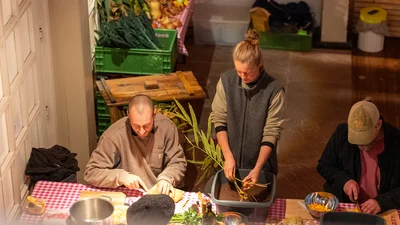 Three people are sitting at a table preparing vegetables, with fruit and vegetable crates in the background.