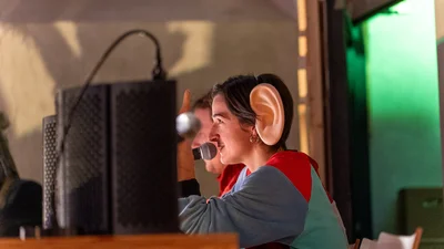 Person with oversized artificial ear sitting at a table with microphone and speakers in the background
