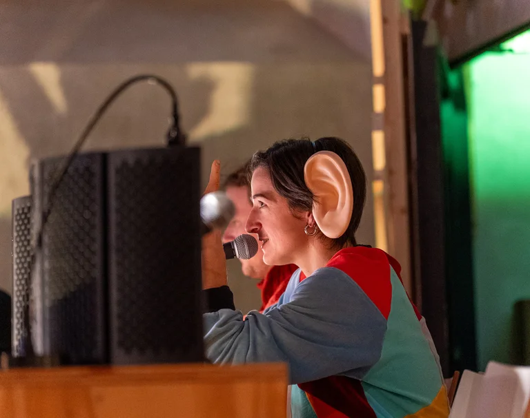 Person with oversized artificial ear sitting at a table with microphone and speakers in the background